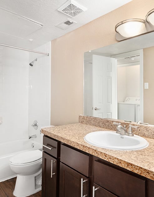 A modern bathroom featuring a granite countertop with dark wood cabinets next to a doorway to bathtub and wood floors at MAA Remington Hills luxury apartments in Irving, TX