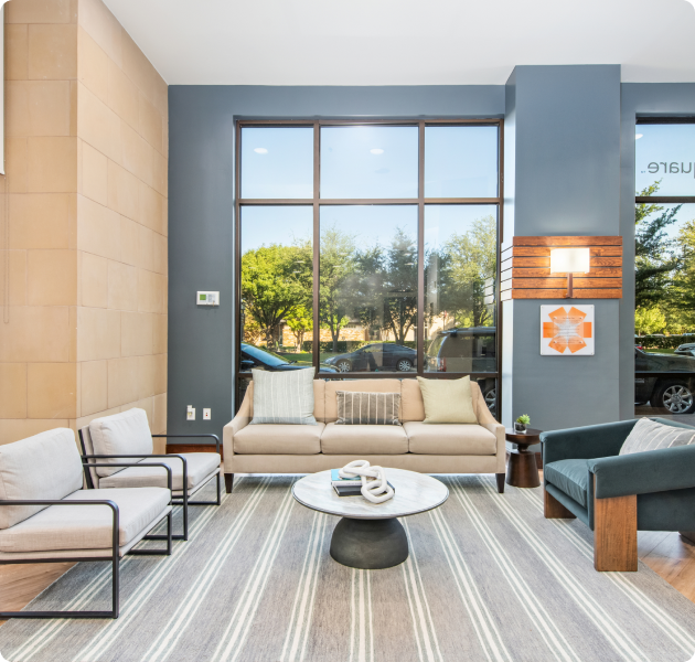 A luxurious community center with a sitting area couch and arm chairs with a circular coffee table, complemented by large picture window views at MAA Times Square luxury apartments in McKinney, TX