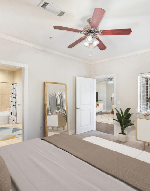 A master bedroom featuring a ceiling fan and a wooden-framed mirror with an ensuite bathroom at MAA Times Square luxury apartments in McKinney, TX