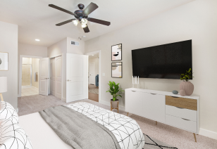 A modern bedroom with a wall-mounted TV, a white dresser with a potted plant, and a ceiling fan at MAA Times Square luxury apartments in McKinney, TX