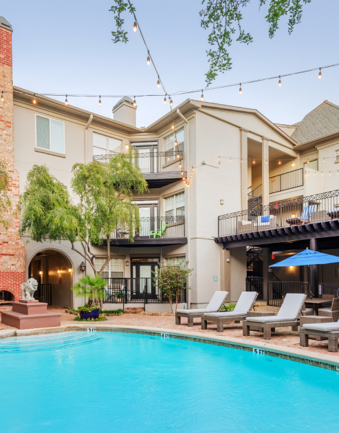 A pool area with lounge chairs and string lights above, surrounded by lush trees, situated next to the apartment complex at MAA Uptown Village luxury apartments in Dallas, TX