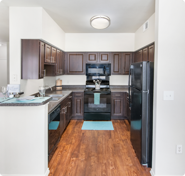 A modern apartment kitchen with dark wood cabinets, black appliances, a double sink and dark gray countertops, with light wood flooring at MAA Watermark luxury apartments in Roanoke, TX