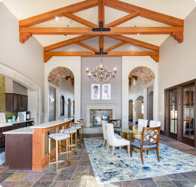 An upscale community center with vaulted ceilings and exposed wooden beams, features seating area with upholstered chairs around a glass dining table, fireplace, bar top counter with bar stools, large arched hallways and patterned rug at MAA Watermark luxury apartments in Roanoke, TX