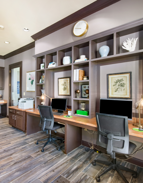 A business center equipped with two workstations, with built-in wooden shelves above the desks displaying various decorations and a wall clock at MAA Watermark luxury apartments in Roanoke, TX