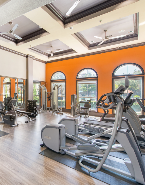 A fitness center equipped with various exercise equipment, mirrored walls, a ceiling fan and large picture windows at MAA Watermark luxury apartments in Roanoke, TX
