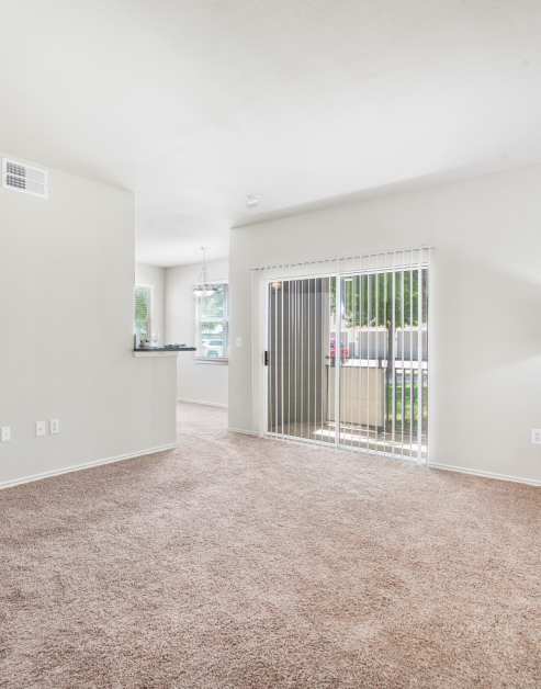 A spacious open living room with beige carpet and large sliding glass doors with vertical blinds adjacent to the kitchen at MAA Watermark luxury apartments in Roanoke, TX