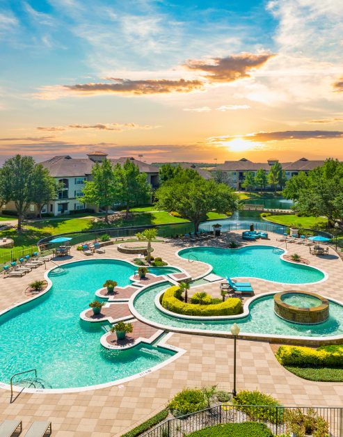 Resort-style pool features wading areas, bordered by lounge chairs and umbrellas, surrounded by lush greenery and trees at MAA Watermark luxury apartments in Roanoke, TX