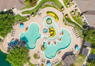 An aerial view of swimming pool with a unique curvy design and multiple wading areas with a central hot tub, surrounded by lounge chairs, umbrellas and lush greenery at MAA Watermark luxury apartments in Roanoke, TX