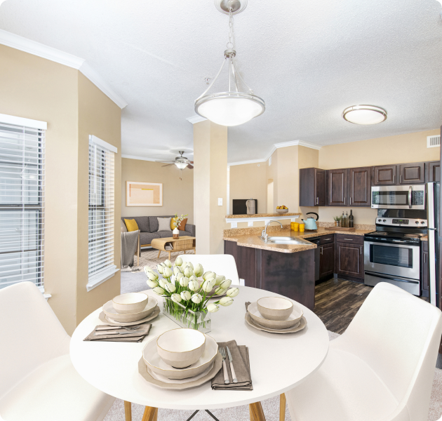 A modern apartment dining area with white round dining table and chairs under a hanging light fixture with bay windows and blinds next to open kitchen at MAA Willow Creek luxury apartments in Bedford, TX