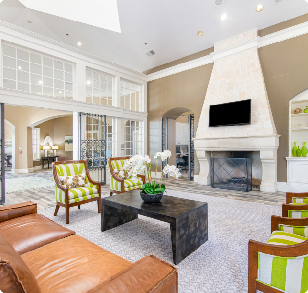 A spacious clubhouse lounge sitting area with a wall-mounted TV above a grand fireplace next to French doors to leasing office at MAA Willow Creek luxury apartments in Bedford, TX