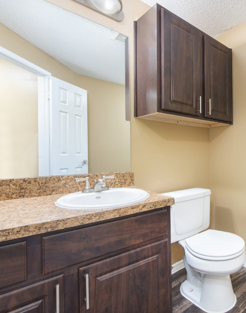 A modern bathroom features spacious cabinets above the toilet and a large vanity mirror and granite countertop with sink and dark wood cabinets at MAA Willow Creek luxury apartments in Bedford, TX