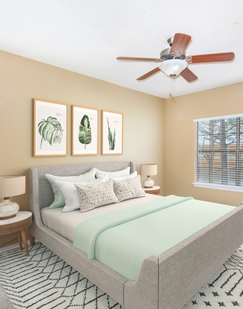 A model guest bedroom with framed wall art above the bed and nightstands, features a ceiling fan and window with blinds at MAA Willow Creek luxury apartments in Bedford, TX