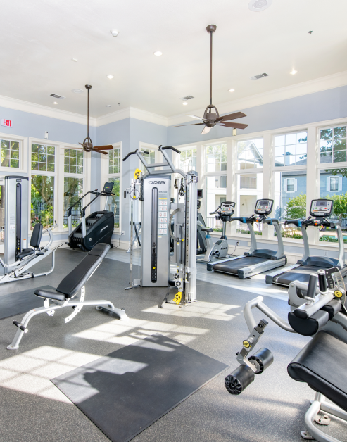 A fitness center with weight machines and treadmills next to large picture windows with ceiling fans at MAA Willow Creek luxury apartments in Bedford, TX