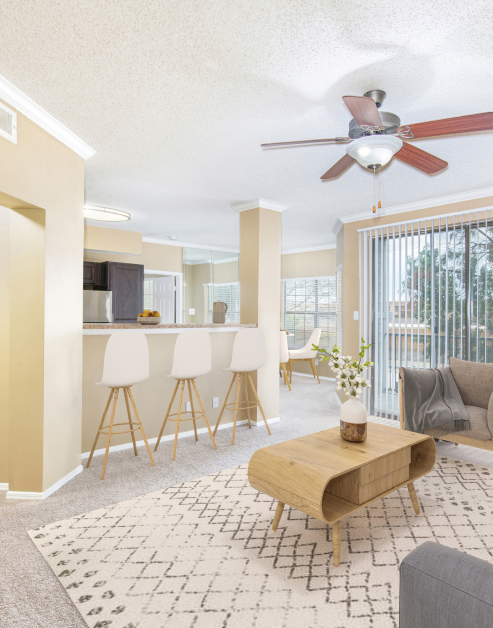 A spacious living room with coffee table and a couch next to kitchen bar top with stools, ceiling fan and sliding glass doors with blinds at MAA Willow Creek luxury apartments in Bedford, TX