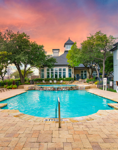 Relaxing two-tiered pool with a waterfall and fountain jets on a stone deck with lounge chairs next to clubhouse, surrounded by lush trees at MAA Willow Creek luxury apartments in Bedford, TX