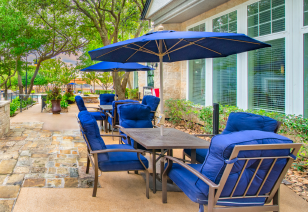 A poolside patio table with blue-cushioned chairs and sun umbrella, next to a stone pathway surrounded by trees at MAA Willow Creek luxury apartments in Bedford, TX