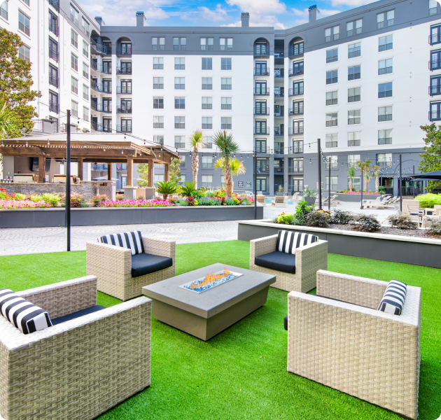 An outdoor seating area with wicker sofas with striped cushions surrounding a modern fire pit, all nestled on a grass lawn surrounded by planters at MAA Worthington luxury apartments in Dallas, TX