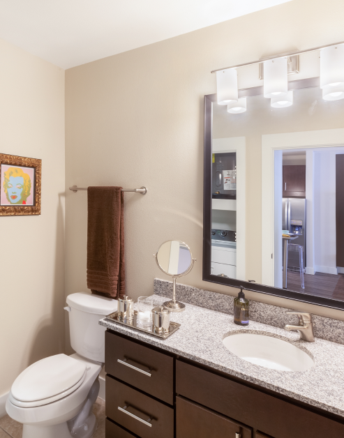 Modern bathroom with light fixtures above large framed vanity mirror with single sink on grey countertop and dark wood cabinets next to toilet at MAA 510 luxury apartments in Houston, TX