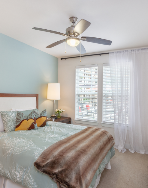 Bedroom with ceiling fan and light blue accent wall with blue bedding and nightstand with lamp next to windows with curtains at MAA 510 luxury apartments in Houston, TX