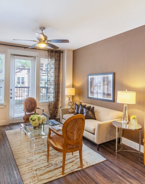 Model living room with warm-toned accent wall with ceiling fan and a French door to balcony next to a sitting area with glass coffee table on an area rug with wood floors at MAA 510 luxury apartments in Houston, TX