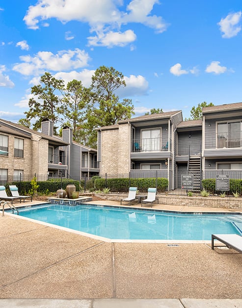 Long view of swimming pool with entrance steps and lounge chairs surrounded by lush greenery next to the apartment complex at MAA Cypresswood luxury apartments in Spring, TX