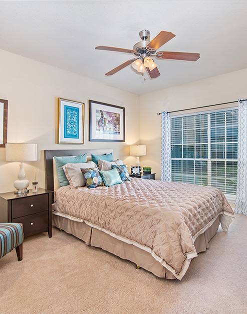 Modern style bedroom with ceiling fan, wall art above bed with ascent pillows, night stands with lamps next to large window with blinds and curtains at MAA Energy Park luxury apartments in Houston, TX