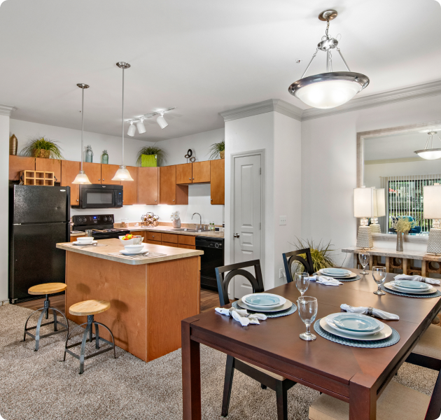 Apartment kitchen with light brown cabinets, upgraded appliances and island countertop next to a dining area with dark wood dining table and chairs at MAA Fall Creek luxury apartments in Humble, TX