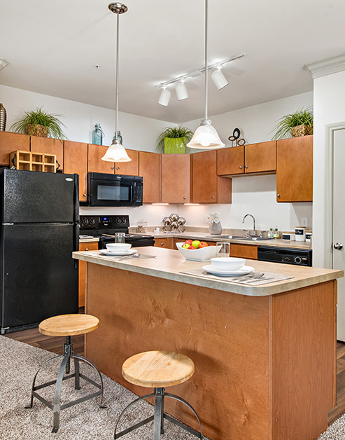 Open-style kitchen with hanging light fixtures, light brown cabinets and upgraded appliances with an island countertop and bar stools on wood floors at MAA Fall Creek luxury apartments in Humble, TX
