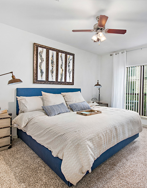 Carpeted bedroom with a ceiling fan, wall art above blue and white bedding, nightstands with lamps next to a window with blinds at MAA Fall Creek luxury apartments in Humble, TX