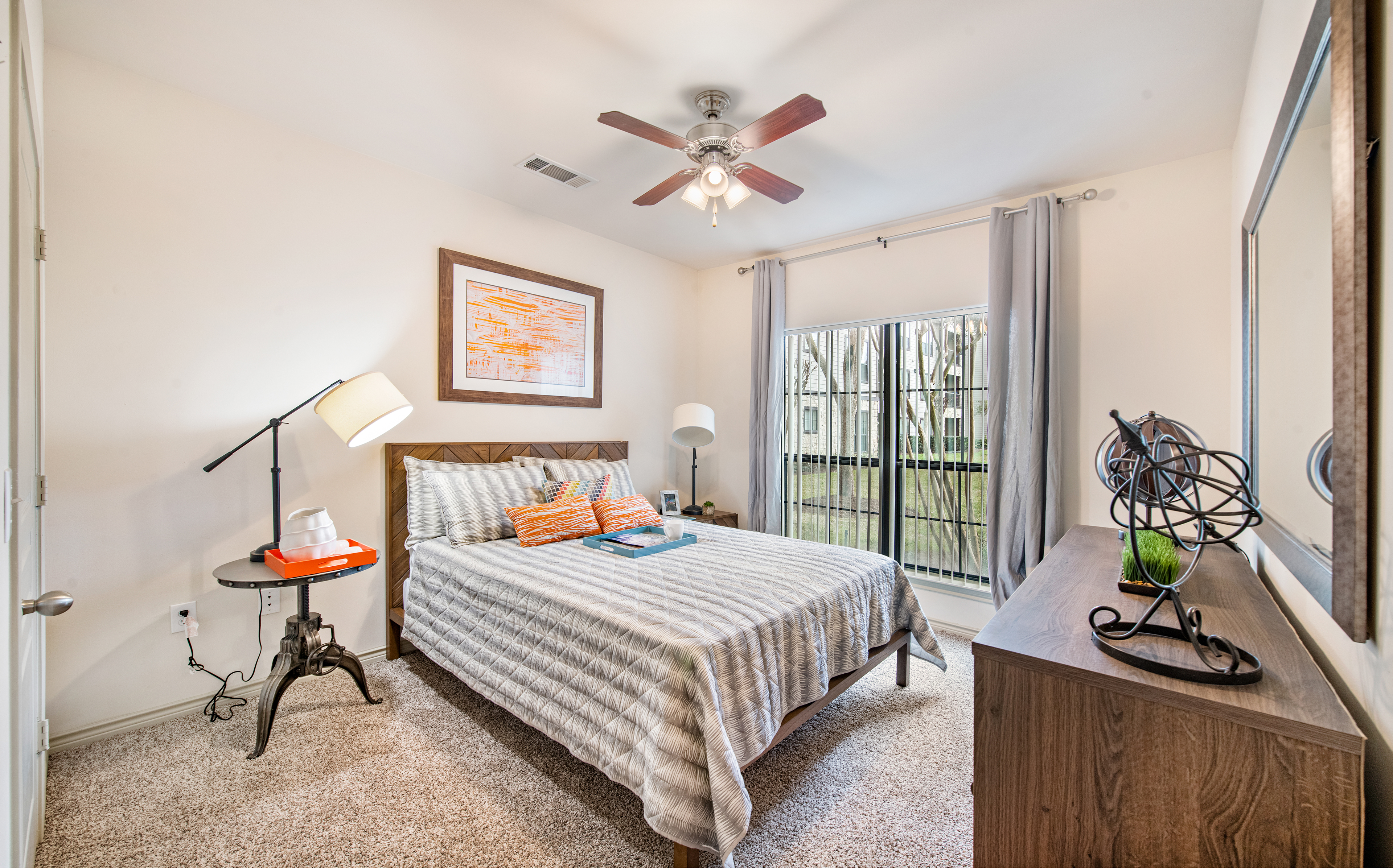A wide view of bedroom with a ceiling fan, wooden dresser and wall art above bed, nightstands with lamps next to a large picture window with blinds and curtains at MAA Fall Creek luxury apartments in Humble, TX