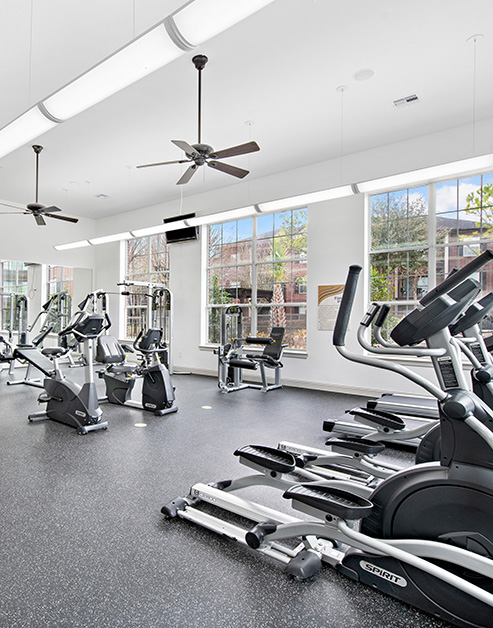 A spacious fitness center with ceiling fans, wall-mounted TV and a variety of workout machines next to large picture windows at MAA Fall Creek luxury apartments in Humble, TX