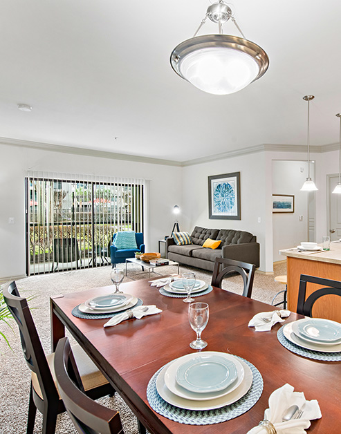 A living room with large picture window with vertical blinds and dark sofa next to a dark wood dining table and chairs at MAA Fall Creek luxury apartments in Humble, TX