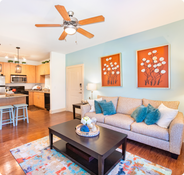 Apartment living area with a ceiling fan, framed orange flower art above couch with pillows and a dark wood coffee table on an area rug and wood floors at MAA Grand Cypress luxury apartments in Cypress, TX