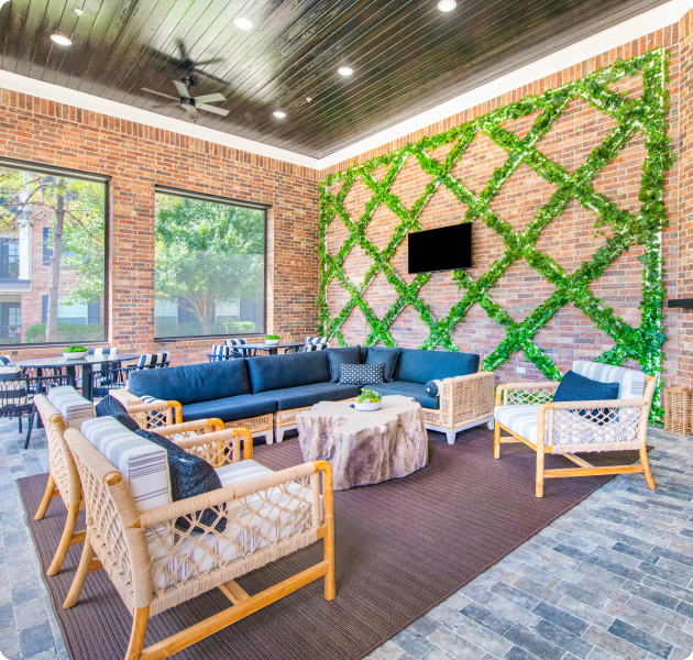 Community screened lanai lounge area enclosed by a brick walls with greenery designs, dark wood plank ceiling with a fan and multiple seating areas on brick flooring at MAA Grand Cypress luxury apartments in Cypress, TX