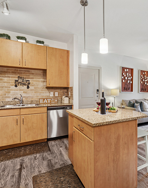 Modern open-style kitchen with hanging light fixtures over island countertop, tan cabinets and brick backsplash with stainless-steel appliances on wood floors at MAA Grand Cypress luxury apartments in Cypress, TX