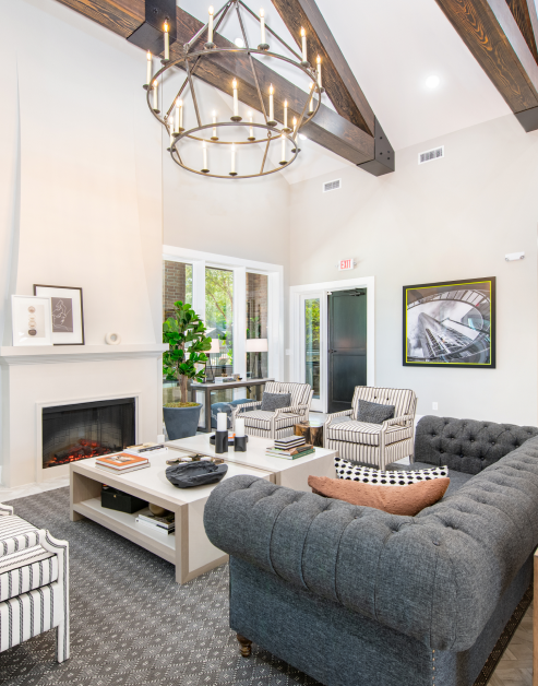 Community clubhouse features a high vaulted ceiling with wood beams and multi-tiered chandelier, modern gray sofa and a light-colored coffee table next to a fireplace at MAA Grand Cypress luxury apartments in Cypress, TX