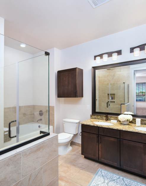Luxury bathroom with light fixtures above a large vanity mirror with double sinks and wooden cabinets along with bathtub and toilet at MAA Greater Heights luxury apartments in Houston, TX