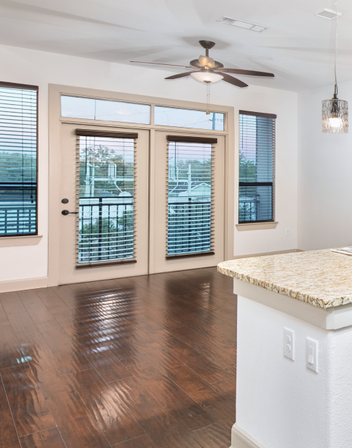 Spacious empty wooden floor living room with a ceiling fan, windows next to glass doors near kitchen area at MAA Greater Heights luxury apartments in Houston, TX