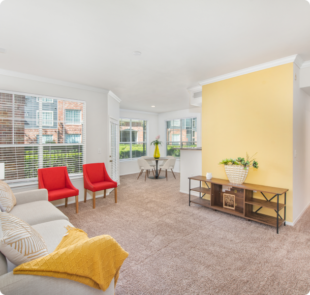 Model apartment living room with a light grey sofa with decorative pillows and two red armchairs, wooden side table with shelves against yellow accent wall at MAA Kirkwood luxury apartments in Stafford, TX