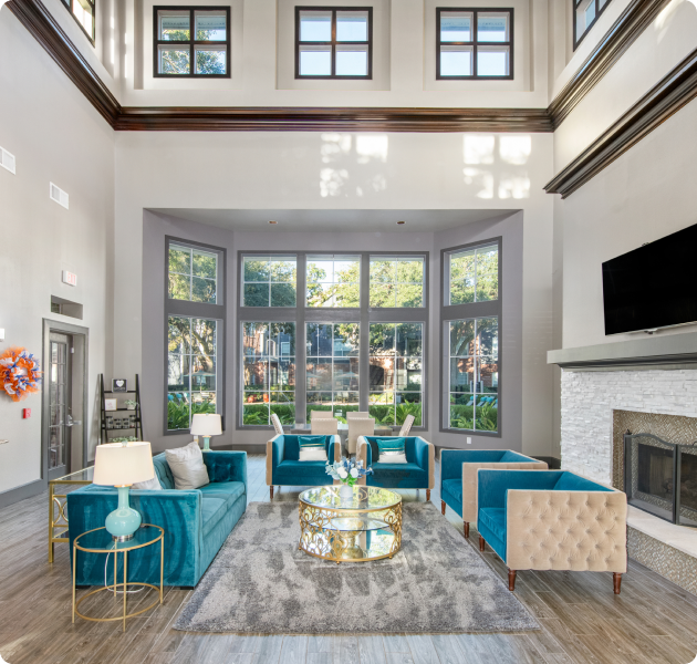Community clubhouse with a high ceiling with large windows, fireplace with a wall-mounted TV next to a seating area with blue armchairs, glass-topped coffee table on an area rug and wood floors at MAA Kirkwood luxury apartments in Stafford, TX