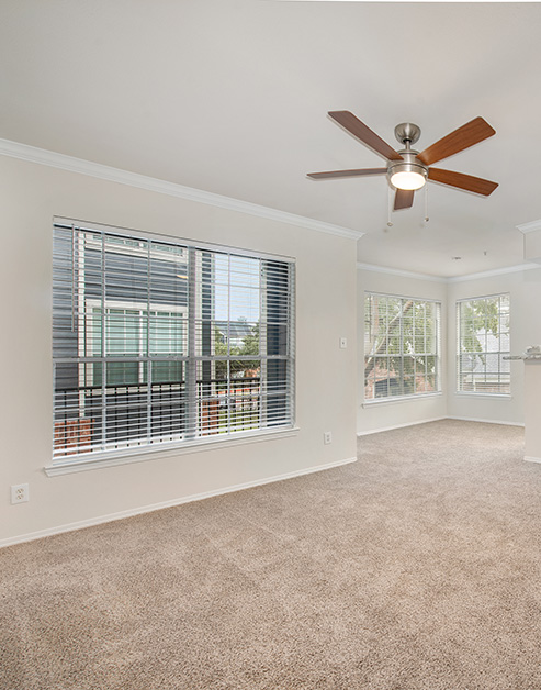 Empty spacious carpeted living room with a ceiling fan, large picture windows with blinds at MAA Kirkwood luxury apartments in Stafford, TX