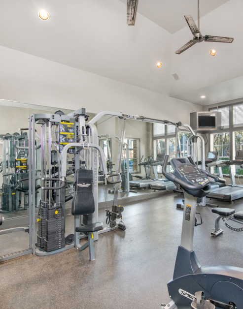 Modern fitness center with ceiling fans, treadmills and weight machines next to a mirrored wall at MAA Kirkwood luxury apartments in Stafford, TX