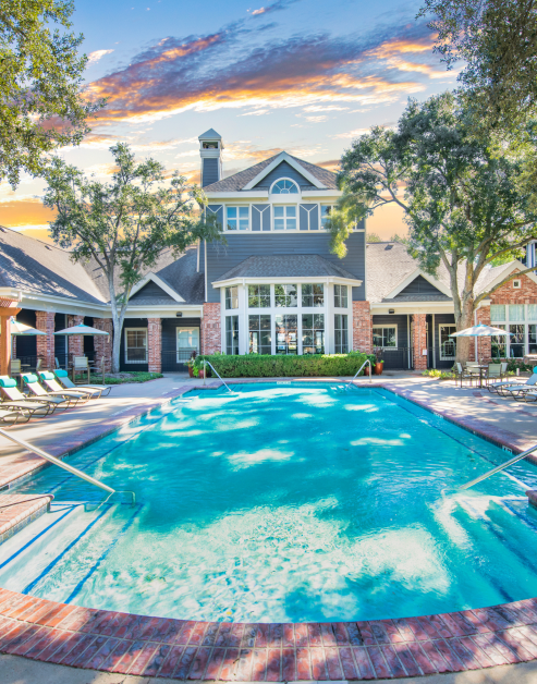 A pool with entrance steps and handrails surrounded by lounge chairs and lush trees next to the clubhouse at MAA Kirkwood luxury apartments in Stafford, TX