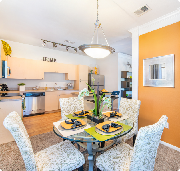 Apartment kitchen and dining area and a hanging light fixture above glass top dining table and chairs next to an orange accent wall at MAA Legacy Pines luxury apartments in Houston, TX