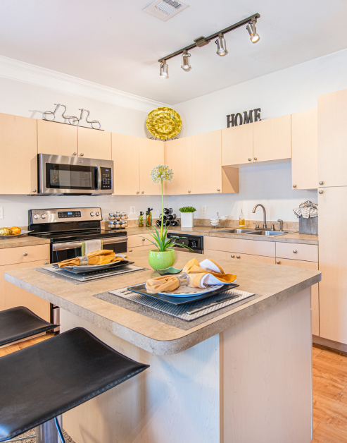Modern-style kitchen with track lighting and island bar with barstool chairs, light tan cabinets with stainless-steel appliances and wood floors at MAA Legacy Pines luxury apartments in Houston, TX