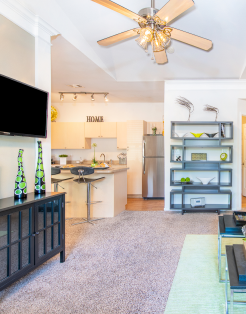 Carpeted living room with ceiling fan, wall-mounted TV above side cabinet with a modern-style bookcase w next to the dining area at MAA Legacy Pines luxury apartments in Houston, TX