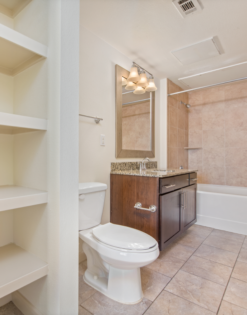 Bathroom with built-in shelving, a framed vanity mirror above granite countertops and dark wood cabinets, tiled shower and bathtub on tiled floors at MAA Midtown Square luxury apartments in Houston, TX
