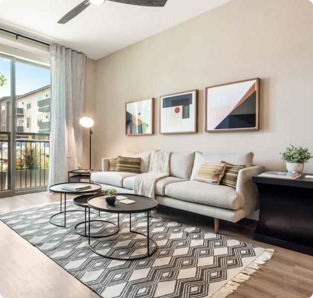 Apartment model living room with wood floors, wall art above white couch with accent pillows, modern-style round coffee tables on an area rug next to glass sliding doors with a balcony view at MAA Park Point luxury apartments in Houston, TX