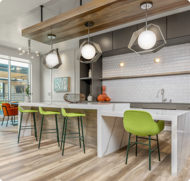 Modern community clubhouse with an open kitchen with white countertop and green chairs and contemporary hanging light fixtures on wooden floors at MAA Park Point luxury apartments in Houston, TX