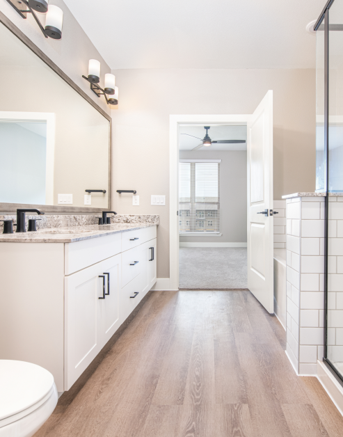 Modern ensuite bathroom with light fixtures above a large vanity mirror with double sinks and white cabinets with wood floors at MAA Park Point luxury apartments in Houston, TX